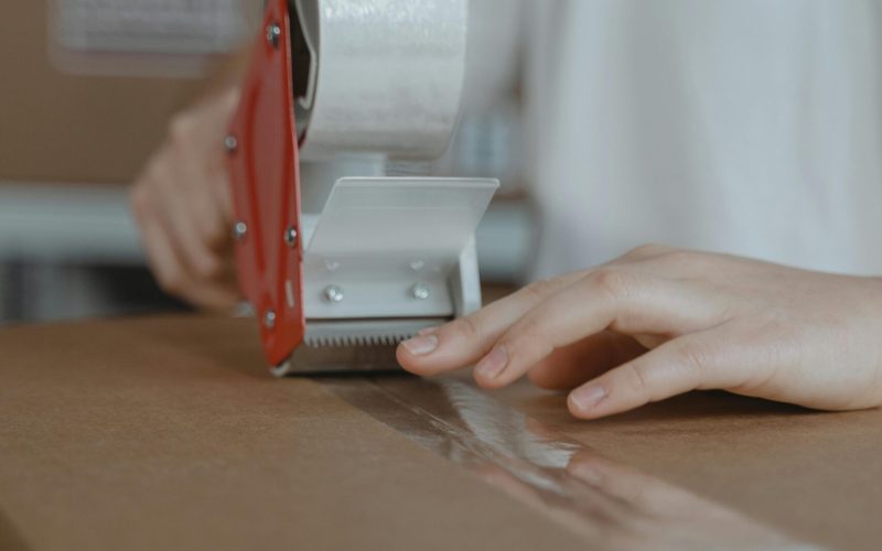 A person sealing a cardboard box with a tape dispenser in an indoor setting.